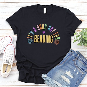May include: Black t-shirt with the text "IT'S A GOOD DAY FOR BEADING" in a colorful, arched design. The design includes a purple heart and floral accents. The shirt is paired with denim shorts and white sneakers.