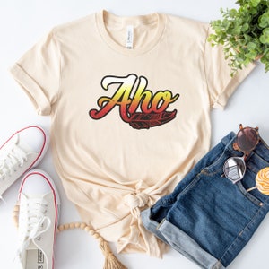 May include: A beige t-shirt with the word "Aho" in a colorful gradient font with a feather design. The shirt is tied in a knot and is laying on a white surface with a pair of white sneakers, a pair of blue denim shorts, and a wooden bead necklace.