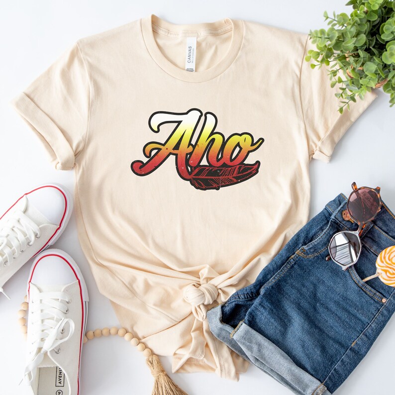 May include: A beige t-shirt with the word "Aho" in a colorful gradient font with a feather design. The shirt is tied in a knot and is laying on a white surface with a pair of white sneakers, a pair of blue denim shorts, and a wooden bead necklace.