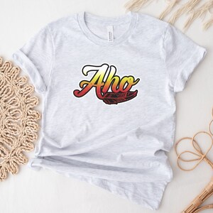 May include: A gray t-shirt with the word "Aho" in a stylized font with a red, orange, and yellow gradient. A black and white feather design is below the word.