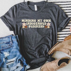 May include: A dark gray t-shirt with the text "Minding My Own Indigenous Business" in a retro font with colorful flowers on either side of the text.