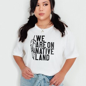 May include: White t-shirt with a black feather design and the text "WE ARE ON NATIVE LAND".