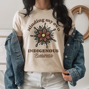 May include: A beige t-shirt with the text "Minding my own Indigenous Business" in brown lettering. A stylized flower with red petals and green leaves is above the text.