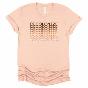 Puede incluir: Una camiseta rosa claro con la palabra "DECOLONIZE" repetida en un degradado de tonos marrones.