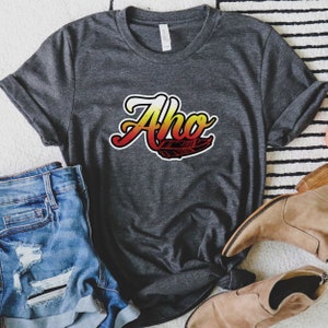 May include: A dark grey t-shirt with the word "Aho" in a stylized font with a red, yellow, and white gradient. A black feather is below the word.