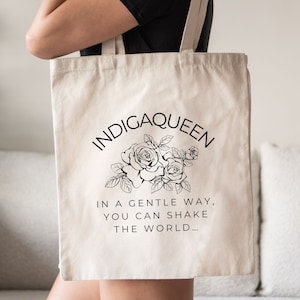 May include: A white canvas tote bag with black text and a floral design. The text reads "Indigqueen" in a circle with roses and leaves. Below the circle, the text reads "In a gentle way, you can shake the world..."