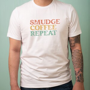 May include: White t-shirt with the text "SMUDGE COFFEE REPEAT" in a vintage style font. The text is in shades of orange, yellow, and green.