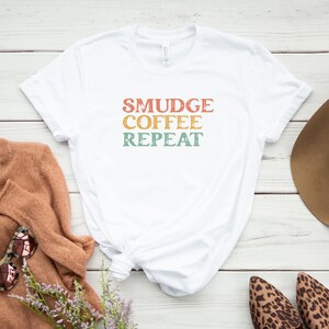 May include: White t-shirt with the text "SMUDGE COFFEE REPEAT" in a retro font with a faded, vintage look. The text is in shades of orange, yellow, and green.