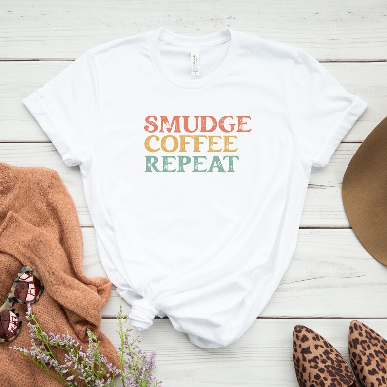 May include: White t-shirt with the text "SMUDGE COFFEE REPEAT" in a retro font with a faded, vintage look. The text is in shades of orange, yellow, and green.