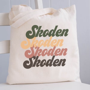 May include: A white canvas tote bag with the word "Skoden" printed four times in a stacked, retro-style font. The text is in shades of green, yellow, pink, and brown.
