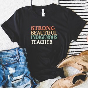 May include: Black t-shirt with the text "STRONG BEAUTIFUL INDIGENOUS TEACHER" in a retro style font with colorful accents.