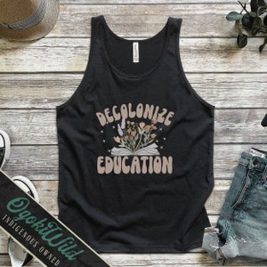 May include: Black tank top with the text "Decolonize Education" in a retro font. The text is surrounded by a floral design with a book underneath the flowers.