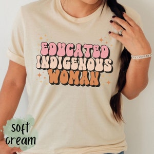 May include: A soft cream colored t-shirt with the text "EDUCATED INDIGENOUS WOMAN" in pink, peach, and brown retro font with gold glitter accents.