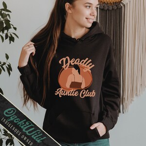 May include: Black hoodie with a brown and orange graphic that says "Deadly Auntie Club". The graphic features a silhouette of a woman with her back turned.