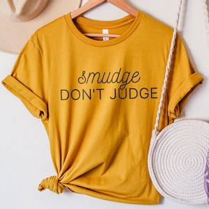 Puede incluir: Una camiseta amarilla mostaza con el texto "smudge don't judge" en letras negras.