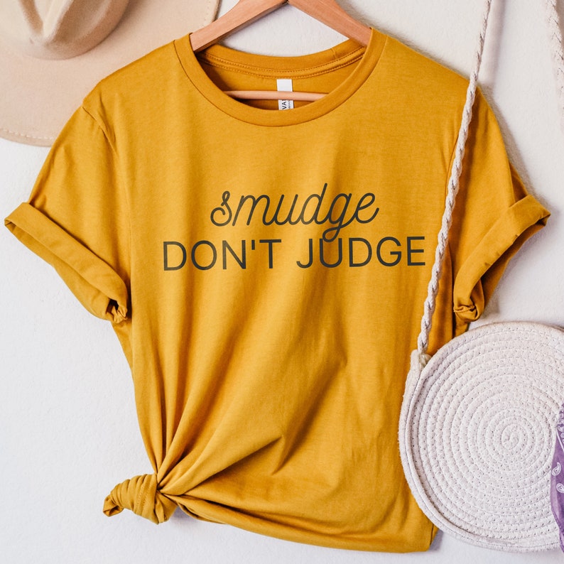 Puede incluir: Una camiseta amarilla mostaza con el texto "smudge don't judge" en letras negras.
