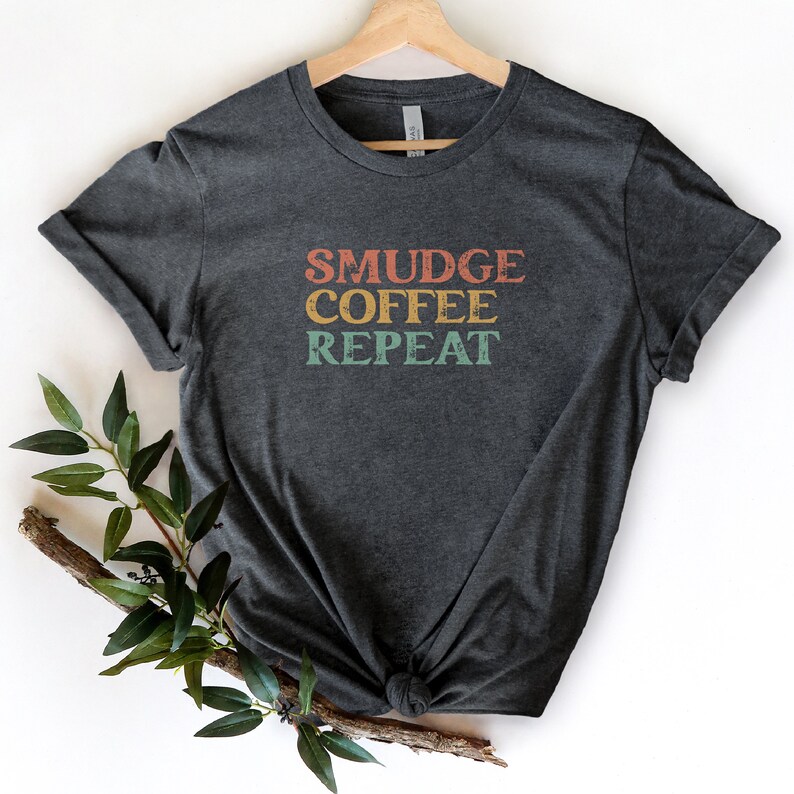 May include: A dark gray t-shirt with the text "SMUDGE COFFEE REPEAT" in a vintage style font with orange, red, and blue colors.