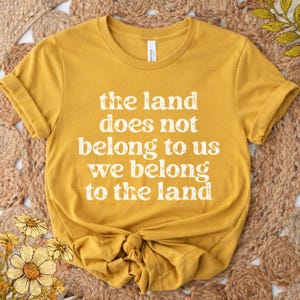 May include: A mustard yellow short-sleeved t-shirt with a crew neckline. The centre of the shirt features white distressed-style text that reads: the land does not belong to us we belong to the land. The shirt has a standard fit and hem.
