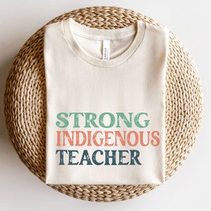 May include: A beige t-shirt with the text "STRONG INDIGENOUS TEACHER" in green, orange, and blue with a distressed look.