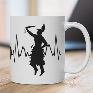 May include: White ceramic mug with a black silhouette of a Native American dancer with a heartbeat line. The dancer is holding a staff and wearing a traditional headdress.
