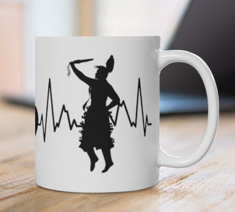 May include: White ceramic mug with a black silhouette of a Native American dancer with a heartbeat line. The dancer is holding a staff and wearing a traditional headdress.