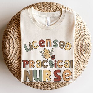 May include: A beige t-shirt with the text "Licensed Practical Nurse" in colorful, retro-style lettering. A cartoon Winnie the Pooh bear is featured in the center of the design.