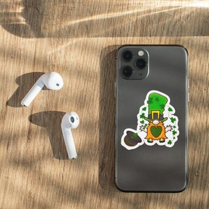 Könnte beinhalten: Ein St. Patrick's Day-Aufkleber mit einem Kobold mit grünem Hut und orangefarbener Weste auf einem Smartphone. Weiße kabellose Ohrhörer liegen auf einer Holzoberfläche.