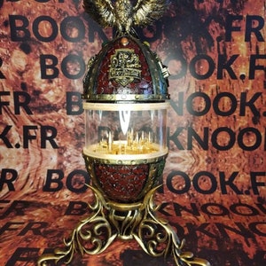 May include: A gold and red steampunk-style lamp with an owl on top and a glass dome containing a miniature city inside. The lamp has a three-legged base with intricate details.