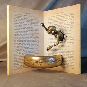 May include: A gold-colored ring with the inscription "One Ring to rule them all, One Ring to find them, One Ring to bring them all and in the darkness bind them" sits on top of an open book. The book is open to a page with text from "The Fellowship of the Ring" by J.R.R. Tolkien. A small, bronze-colored figure of Gollum is emerging from the page.