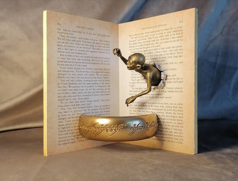 May include: A gold-colored ring with the inscription "One Ring to rule them all, One Ring to find them, One Ring to bring them all and in the darkness bind them" sits on top of an open book. The book is open to a page with text from "The Fellowship of the Ring" by J.R.R. Tolkien. A small, bronze-colored figure of Gollum is emerging from the page.