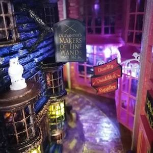 Puede incluir: Un modelo en miniatura de una tienda con un letrero que dice "Ollivander's Makers of Fine Wands Since 382 BC". La tienda est&aacute; iluminada con luces amarillas c&aacute;lidas y tiene un exterior azul oscuro. Un letrero para "Quality Quidditch Supplies" es visible en el fondo.