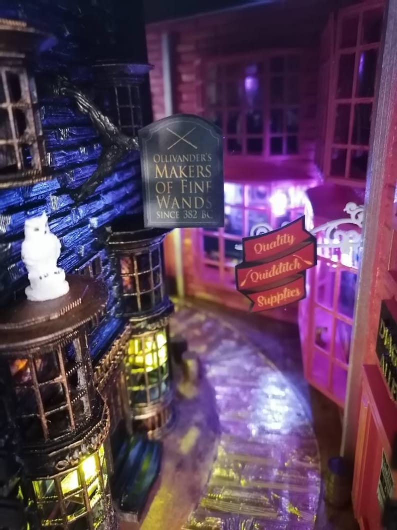Puede incluir: Un modelo en miniatura de una tienda con un letrero que dice "Ollivander's Makers of Fine Wands Since 382 BC". La tienda est&aacute; iluminada con luces amarillas c&aacute;lidas y tiene un exterior azul oscuro. Un letrero para "Quality Quidditch Supplies" es visible en el fondo.