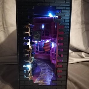 Puede incluir: Un diorama en miniatura de un edificio de ladrillo con un escaparate que dice "Ollivanders Makers of Fine Wands" y un letrero que dice "Leaky Cauldron". El diorama est&aacute; iluminado con luces azules y blancas.