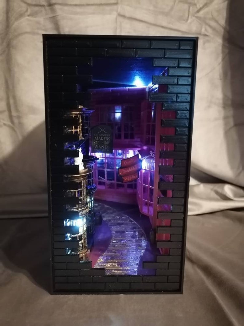 Puede incluir: Un diorama en miniatura de un edificio de ladrillo con un escaparate que dice "Ollivanders Makers of Fine Wands" y un letrero que dice "Leaky Cauldron". El diorama est&aacute; iluminado con luces azules y blancas.