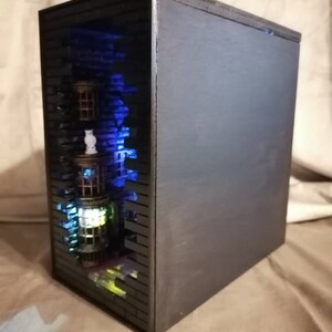 Puede incluir: Una caja de madera negra con un peque&ntilde;o modelo detallado de un edificio en su interior. El edificio est&aacute; iluminado con luces azules y verdes, y la caja est&aacute; abierta por un lado para revelar el modelo.