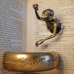 May include: A bronze Gollum figurine emerging from a torn page of a book. The book is open to a page with text about the fictional character Gollum from the Lord of the Rings. The figurine is holding a golden ring with the inscription "One Ring to rule them all" in Elvish script.
