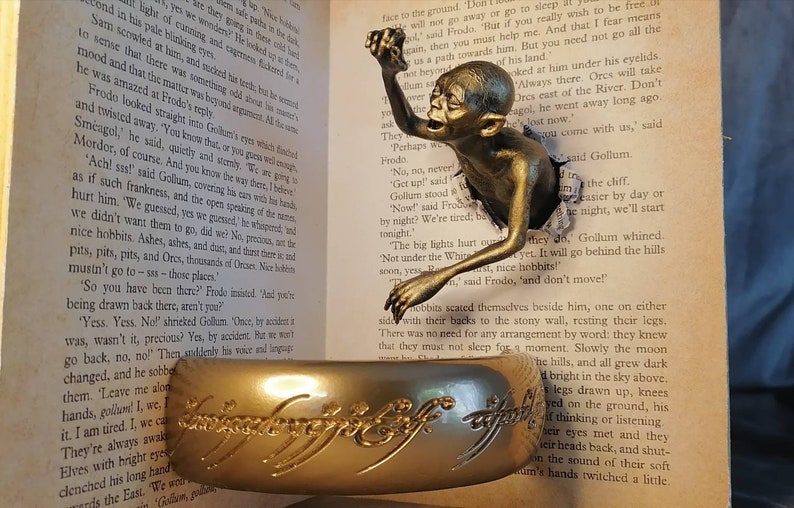 May include: A bronze Gollum figurine emerging from a torn page of a book. The book is open to a page with text about the fictional character Gollum from the Lord of the Rings. The figurine is holding a golden ring with the inscription "One Ring to rule them all" in Elvish script.
