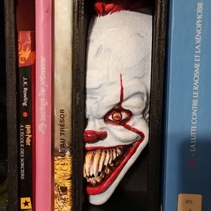 Peut inclure: Un serre-livres représentant un visage de clown peint avec des cheveux roux, une peau blanche et un large sourire aux dents. Le visage du clown est partiellement caché derrière un cadre noir, et le serre-livres est placé sur une étagère avec d'autres livres.