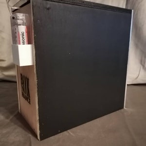 Puede incluir: Una caja de madera negra con las palabras "Book Balance" impresas en el lateral. Un peque&ntilde;o recipiente de pl&aacute;stico blanco con una etiqueta roja y blanca que dice "Isotonic" est&aacute; unido al lateral de la caja.