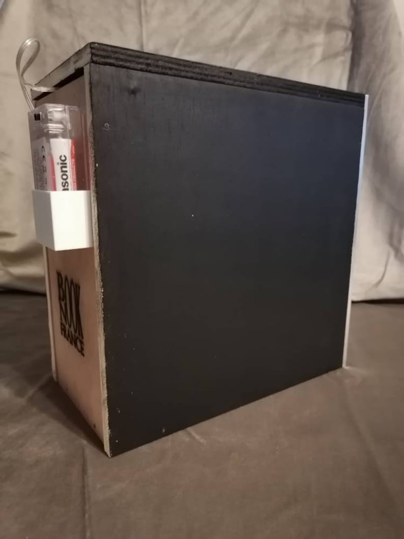 Puede incluir: Una caja de madera negra con las palabras "Book Balance" impresas en el lateral. Un peque&ntilde;o recipiente de pl&aacute;stico blanco con una etiqueta roja y blanca que dice "Isotonic" est&aacute; unido al lateral de la caja.
