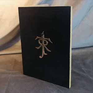 May include: A black book with a silver embossed symbol on the front cover. The symbol is a stylized letter "R" with a cross above it.