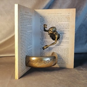 May include: A bronze Gollum figurine from The Lord of the Rings is peeking out of an open book. The book is titled "The Taming of Smaug" and the figurine is holding the One Ring.