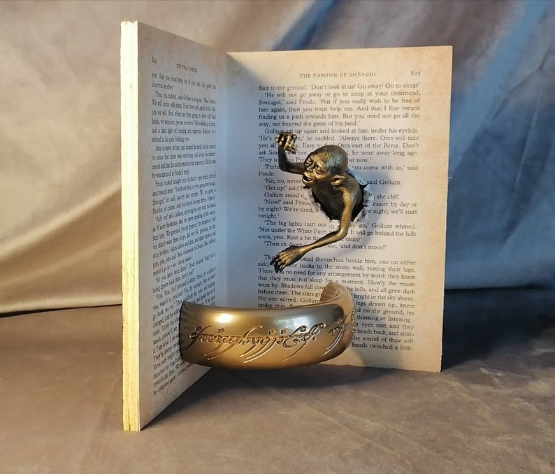May include: A bronze Gollum figurine from The Lord of the Rings is peeking out of an open book. The book is titled "The Taming of Smaug" and the figurine is holding the One Ring.