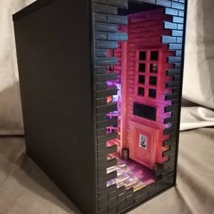 Puede incluir: Una caja negra con una fachada de pared de ladrillo. Un peque&ntilde;o edificio rosa con una puerta y ventanas es visible a trav&eacute;s de un agujero en la pared. El edificio est&aacute; iluminado con luces rosas y moradas.