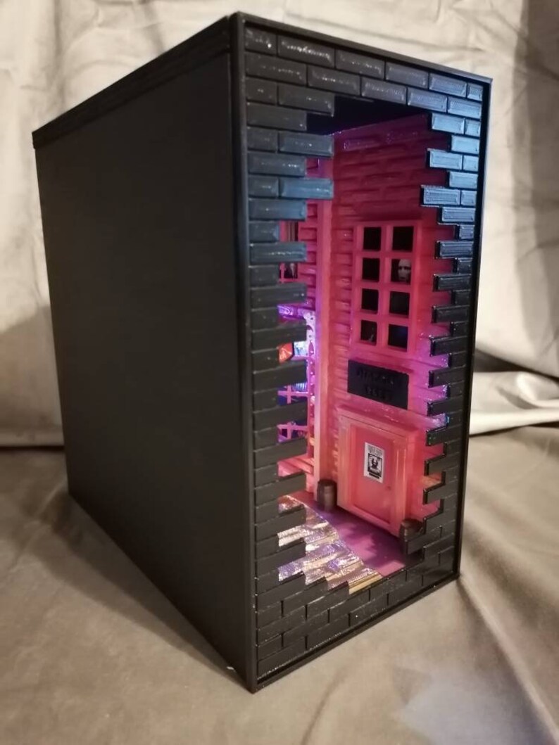 Puede incluir: Una caja negra con una fachada de pared de ladrillo. Un peque&ntilde;o edificio rosa con una puerta y ventanas es visible a trav&eacute;s de un agujero en la pared. El edificio est&aacute; iluminado con luces rosas y moradas.