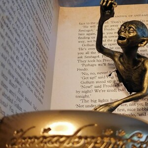 May include: A bronze Gollum figurine from the Lord of the Rings, peeking out of a book with the text "AMING OF SMEAGOL" visible. The figurine is holding a golden ring.