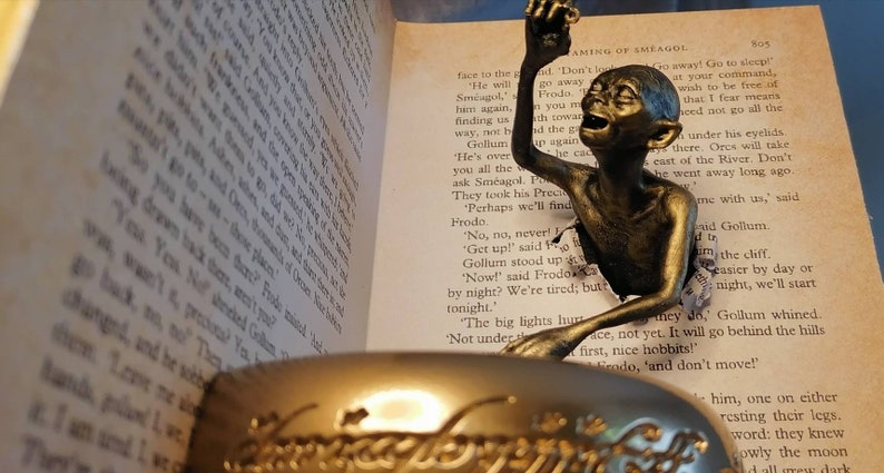 May include: A bronze Gollum figurine from the Lord of the Rings, peeking out of a book with the text "AMING OF SMEAGOL" visible. The figurine is holding a golden ring.