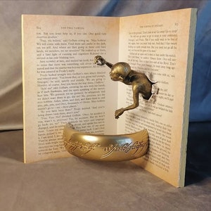 May include: A gold-colored ring with the inscription "The One Ring" is displayed on a book with a bronze-colored figure emerging from the pages. The book is open to a page with text from "The Two Towers" by J.R.R. Tolkien.