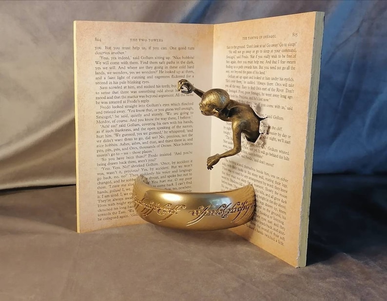 May include: A gold-colored ring with the inscription "The One Ring" is displayed on a book with a bronze-colored figure emerging from the pages. The book is open to a page with text from "The Two Towers" by J.R.R. Tolkien.