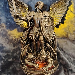 May include: A bronze-toned statue of an archangel with large wings, wearing armor and holding a sword in one hand and a shield with a cross in the other. The figure stands atop a horned creature, with warm orange light glowing from the base.